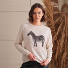 A woman wearing a light beige crewneck sweater with a black and white zebra graphic on the front, standing in front of a neutral wall with dried pampas grass.