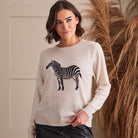 A woman wearing a light beige knit sweater with a black and white zebra graphic on the front, paired with black pants, standing in front of a neutral wall with dried pampas grass.
