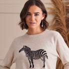 A woman wearing a light beige crewneck sweater with a black and white zebra graphic on the front, posed against a neutral indoor background with dried pampas grass.