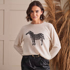 A woman wearing a light beige knit sweater with a black and white zebra graphic on the front, paired with black pants, standing in front of a neutral wall with dried pampas grass.