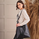 A woman wearing a light beige knit sweater with a black and white zebra graphic on the front, paired with black pants, and large leather slouch bag standing in front of a neutral wall with dried pampas grass.