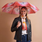 A woman holding a pink umbrella with bird illustrations, wearing a black leather jacket, white t-shirt, and blue jeans against a solid beige background.