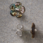 A pair of flower-shaped stud earrings on a textured gray background. One earring is shown from the front, featuring iridescent brown and green petals with a silver center.