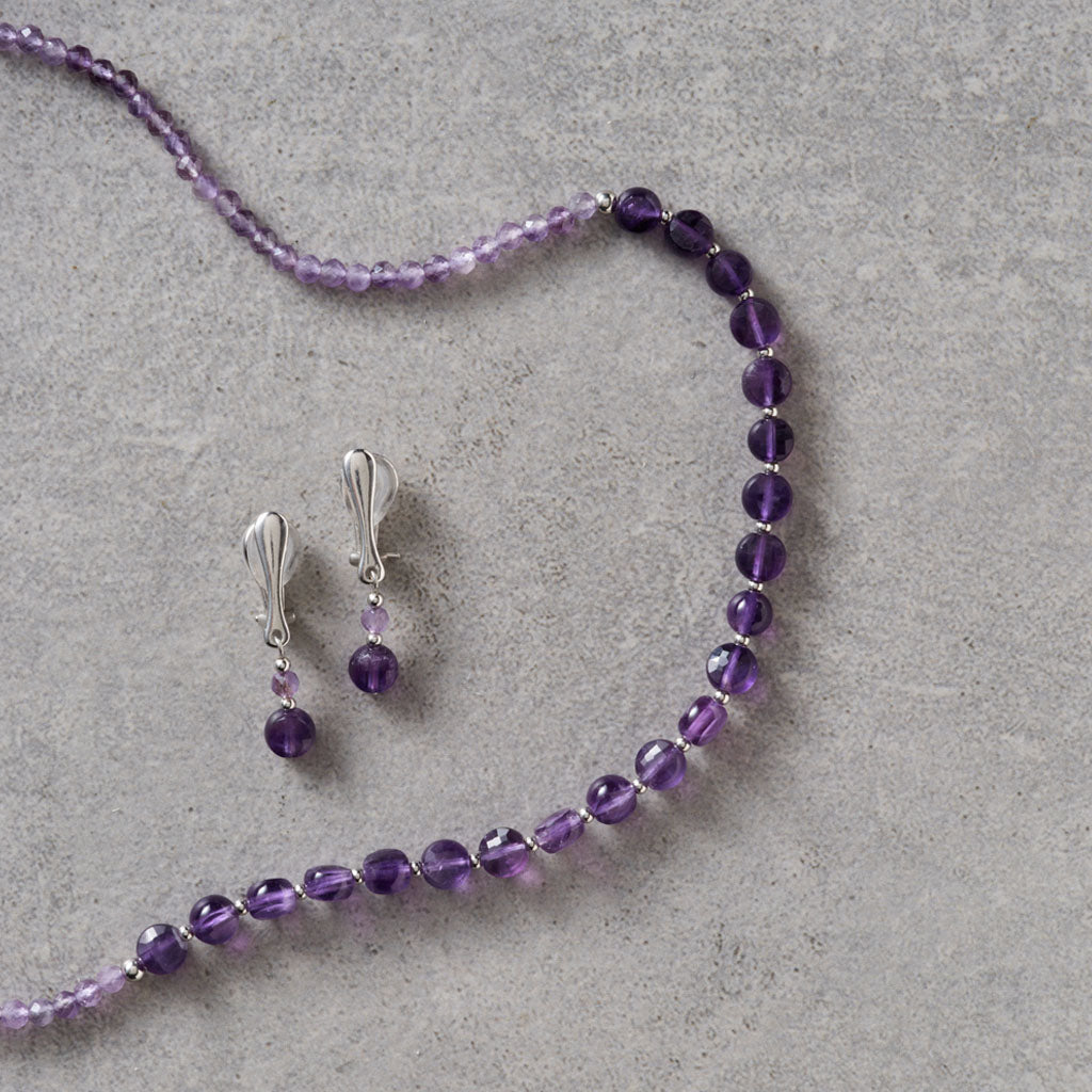 A jewelry set featuring a beaded necklace and matching drop earrings in purple beads with silver-toned metal findings, arranged on a textured gray surface.