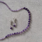 A jewelry set featuring a beaded necklace and matching drop earrings in purple beads with silver-toned metal findings, arranged on a textured gray surface.