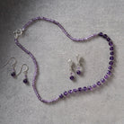 A jewelry set featuring a beaded necklace and matching drop earrings in shades of purple, accented with silver-tone metal findings, displayed on a textured gray background.
