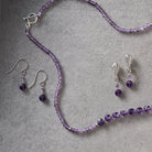 A jewellery set featuring a beaded necklace and matching drop earrings in purple beads with silver-toned metal findings, arranged on a textured gray surface.