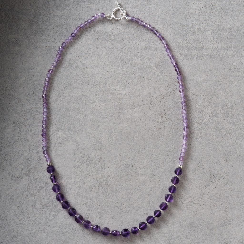 A beaded necklace made of faceted purple beads, with a silver clasp, displayed on a textured gray background.