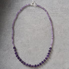 A beaded necklace made of faceted purple beads, with a silver clasp, displayed on a textured gray background.