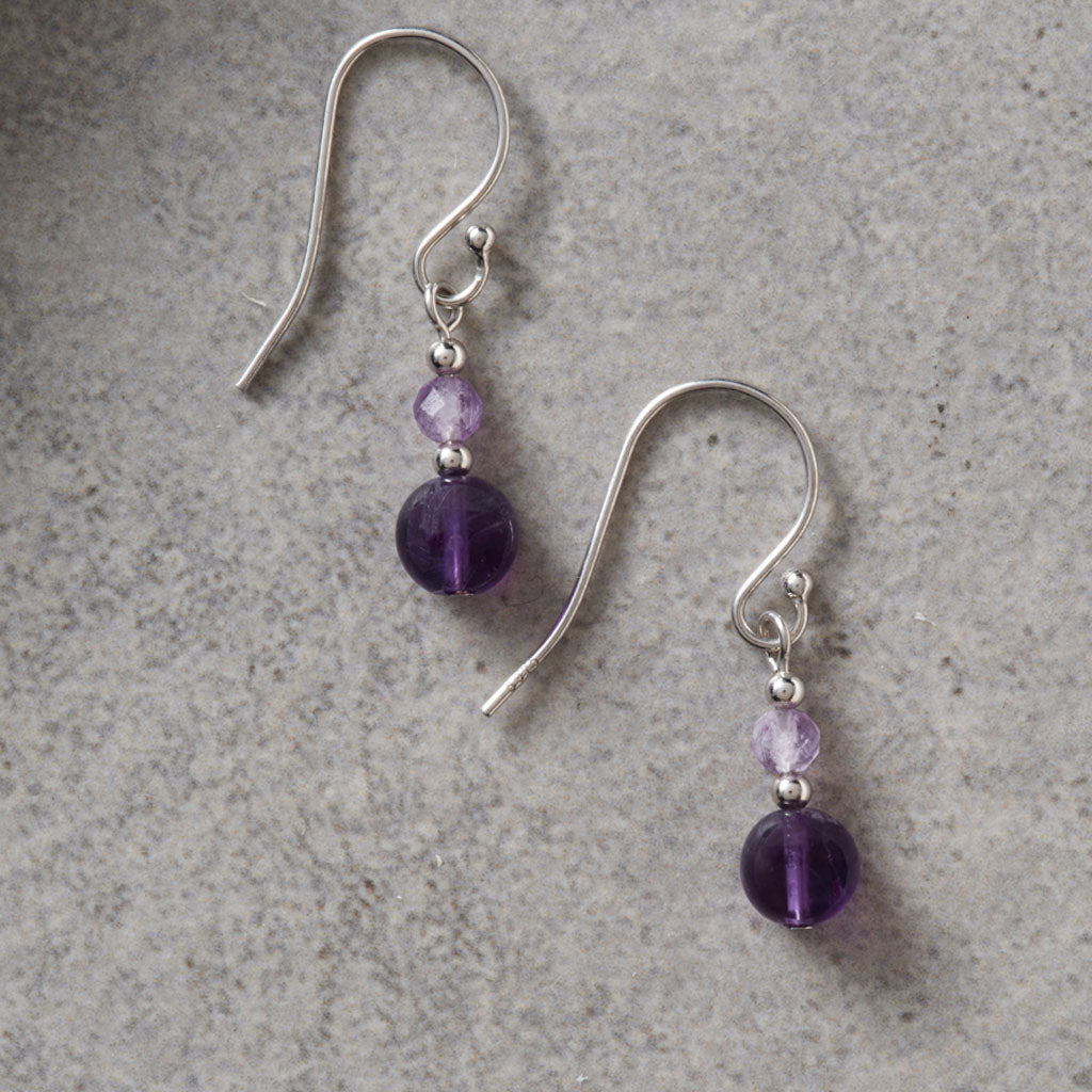 A pair of silver fishhook drop earrings with small faceted purple beads and larger round purple beads, displayed on a textured gray surface.