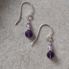 A pair of silver fishhook drop earrings with small faceted purple beads and larger round purple beads, displayed on a textured gray surface.
