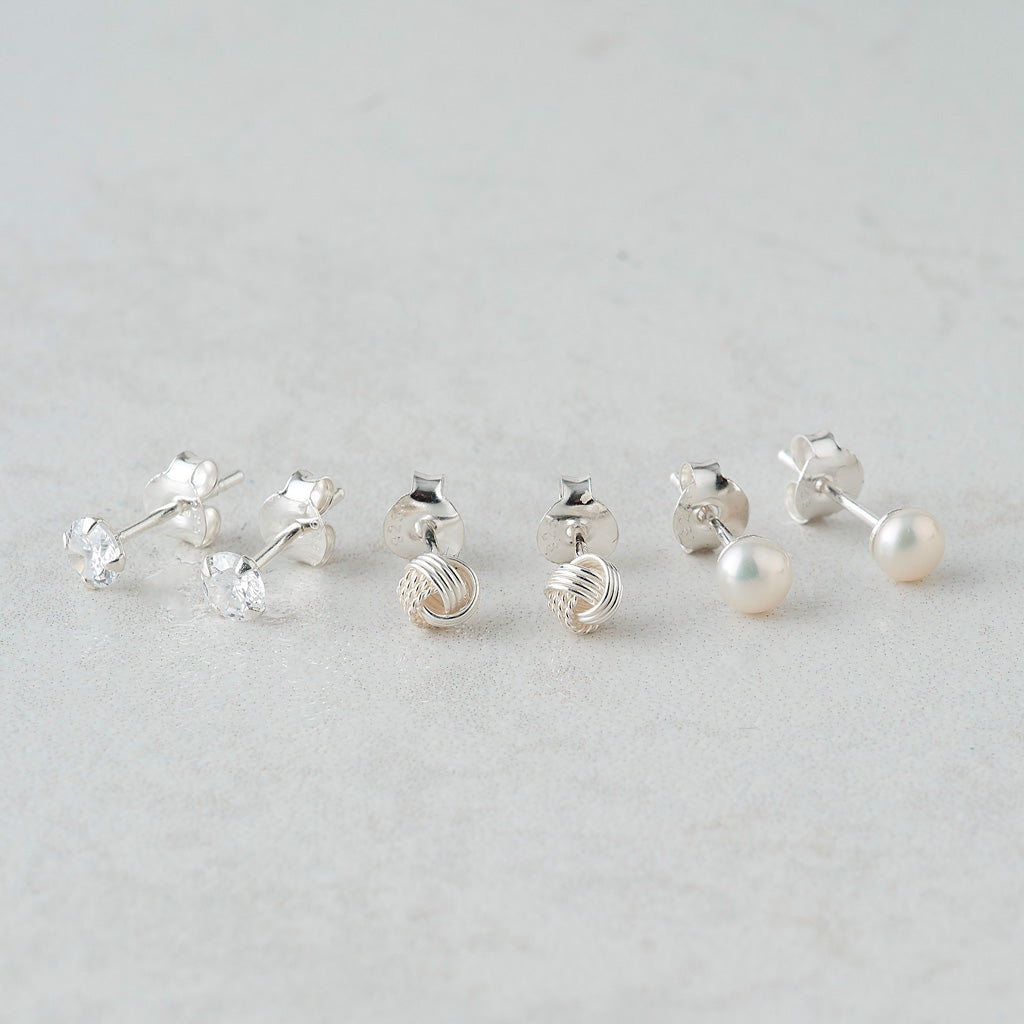 Six pairs of silver stud earrings arranged on a light gray marble surface. The earrings include clear crystal clusters, textured silver ball studs, and white freshwater pearls, showing both front and back views.