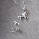 A silver unicorn pendant necklace with a matching stud earring, displayed on a textured gray background. The unicorn pendants feature polished silver bodies with small gold accents on the horns.