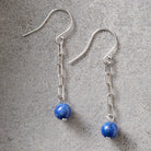 A pair of silver dangle earrings with paperclip chain links and round blue lapis lazuli beads, displayed on a textured gray surface.
