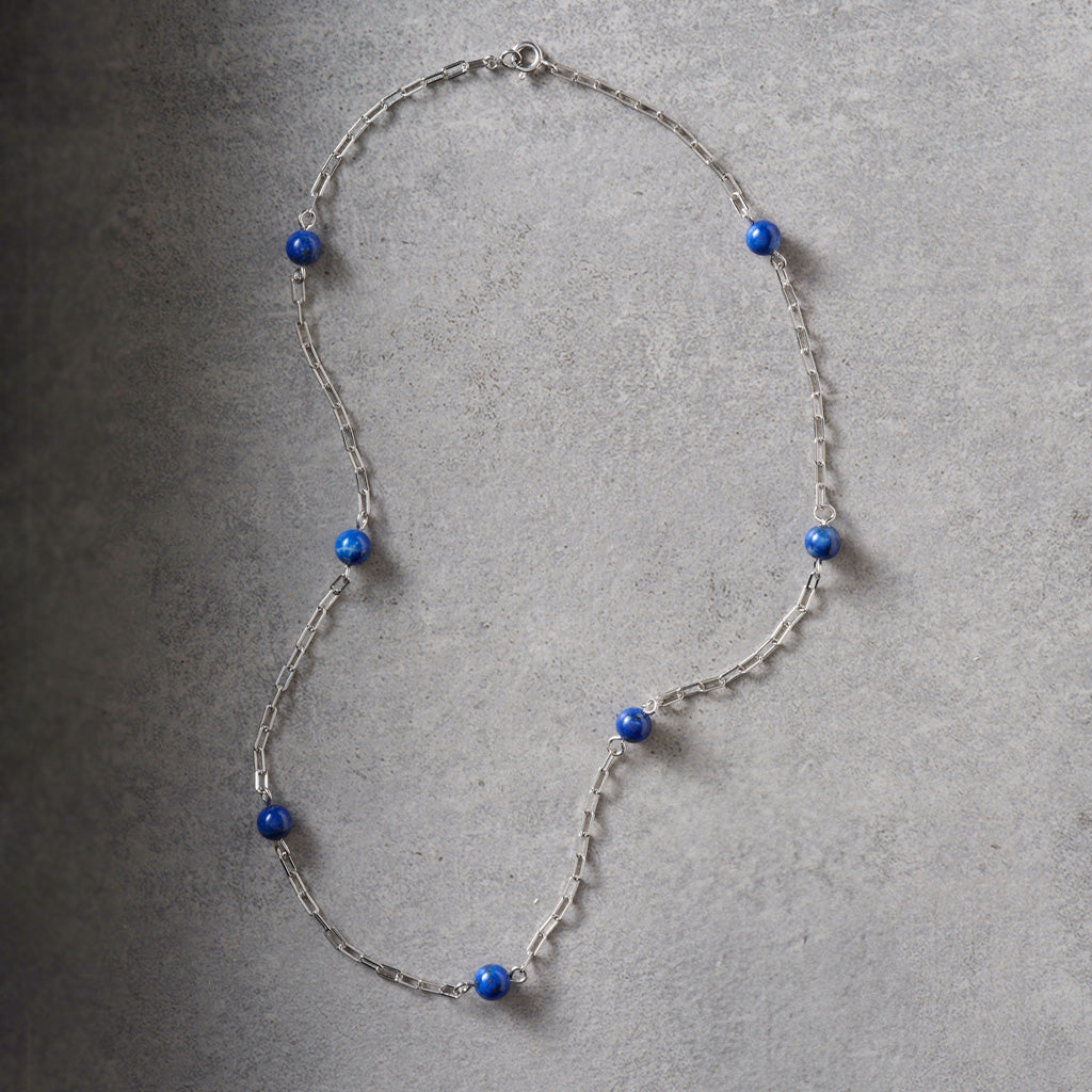 A silver-toned chain necklace with multiple small, round, deep blue beads spaced along the length of the necklace, displayed on a textured gray surface.