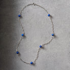 A silver-toned chain necklace with multiple small, round, deep blue beads spaced along the length of the necklace, displayed on a textured gray surface.