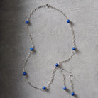 A silver-toned chain necklace and matching drop earrings with small round blue beads, arranged on a textured gray surface.