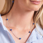 A close-up of a woman wearing a delicate silver chain necklace with five small, round blue beads spaced along the chain. She is also wearing a matching pair of drop earrings, and a light blue and white striped shirt is visible.