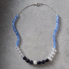 A beaded necklace with faceted light blue beads, clear beads, dark navy beads, and a central clear crystal cluster, displayed on a textured gray background.