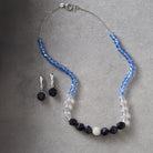 A beaded necklace and matching drop earrings laid flat on a textured gray surface. The necklace has faceted light blue beads, clear beads, dark navy faceted beads, and a central silver-toned round bead with clear stones.