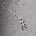 A silver necklace featuring a teddy bear pendant covered in small clear stones, holding a small silver cross charm, displayed on a textured gray surface.