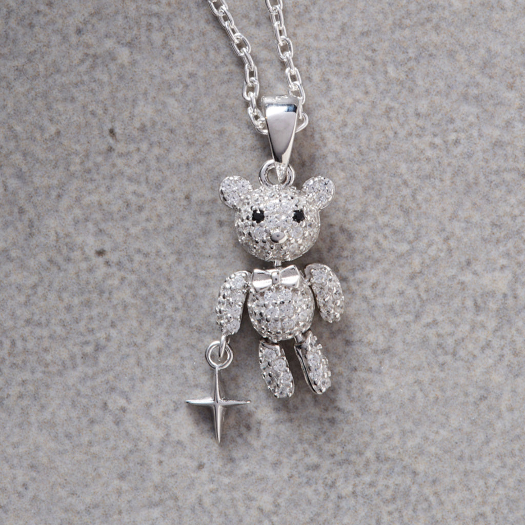 A silver teddy bear pendant necklace encrusted with clear crystals, featuring a small dangling star charm, displayed on a textured gray background.