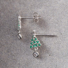 A pair of silver dangle earrings on a textured gray background. One earring is shown from the back with a post and butterfly clutch, and the other is shown from the front with a silver tree-shaped setting paved with green stones and a dangling clear round gemstone.