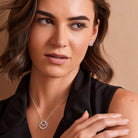 A close-up of a woman wearing a delicate silver necklace with a double-ring pendant and a matching stud earring, set against a warm beige background.