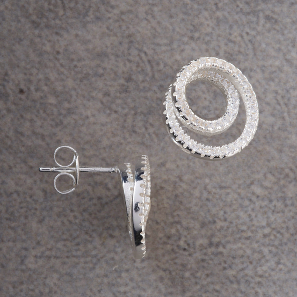 A pair of silver-toned spiral hoop earrings with clear pavé stones, shown on a textured gray background. One earring is displayed from the front showing the spiral design, and the other is turned sideways to show the post and butterfly back.