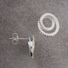 A pair of silver-toned spiral hoop earrings with clear pavé stones, shown on a textured gray background. One earring is displayed from the front showing the spiral design, and the other is turned sideways to show the post and butterfly back.