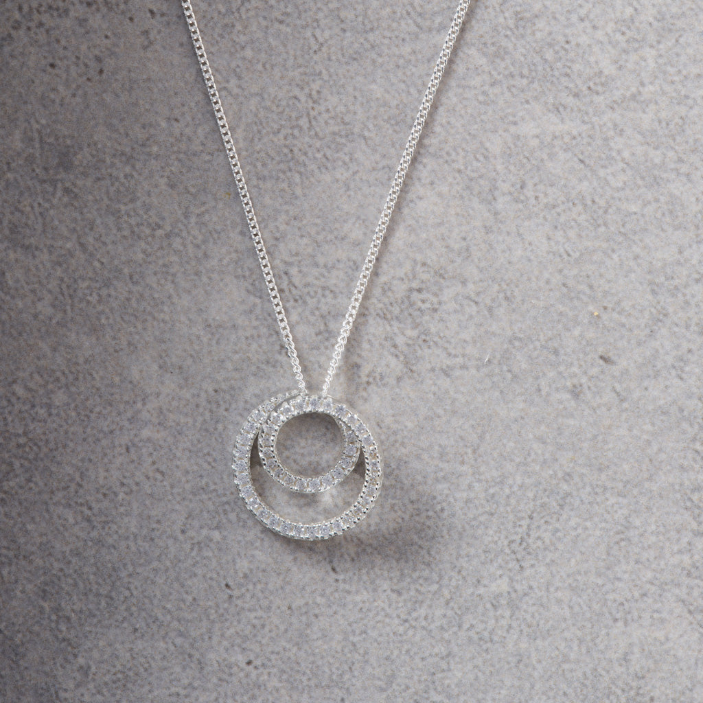 A silver necklace with a double-ring pendant paved with small clear stones, displayed on a textured gray surface.