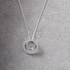 A silver necklace with a double-ring pendant paved with small clear stones, displayed on a textured gray surface.