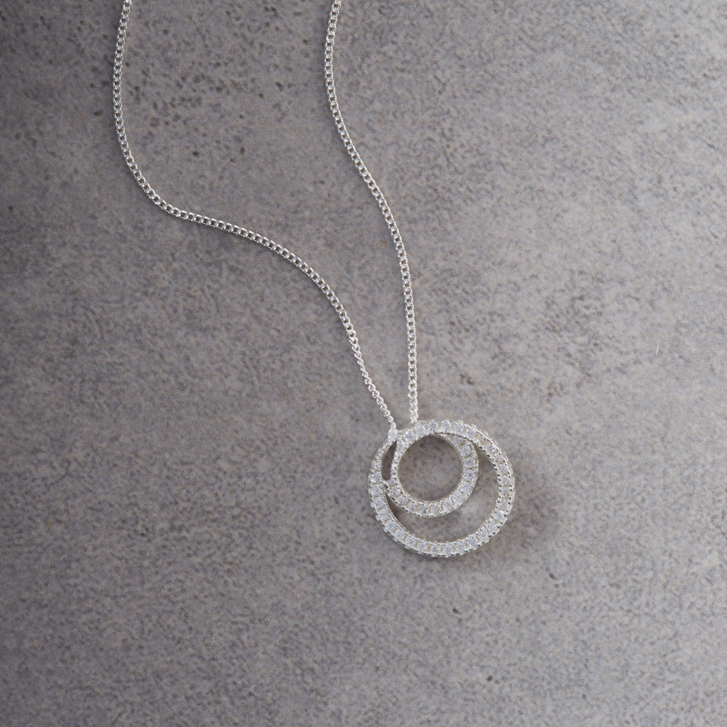 A silver chain necklace with a double-ring pendant paved with small clear stones, displayed on a textured gray surface.