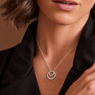 A close-up of a model wearing a delicate silver necklace with a double-ring pendant adorned with small clear stones, styled with a black satin shirt against a warm brown background.