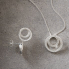 Silver-tone jewelry set featuring a necklace with a double-ring pendant and matching stud earrings, displayed on a textured gray surface.