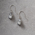 A pair of silver drop earrings with a small clear faceted bead and a square silver charm, displayed on a textured gray surface.