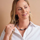 A woman with blonde hair wearing a beaded necklace and matching earrings, posing in a white collared shirt against a solid beige background.