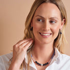 A smiling blonde woman wearing a beaded necklace and matching drop earrings, photographed against a warm beige background.
