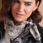 A close-up of a woman wearing a black leather jacket and a cream scarf with a dark brown map-like print and dark purple border.