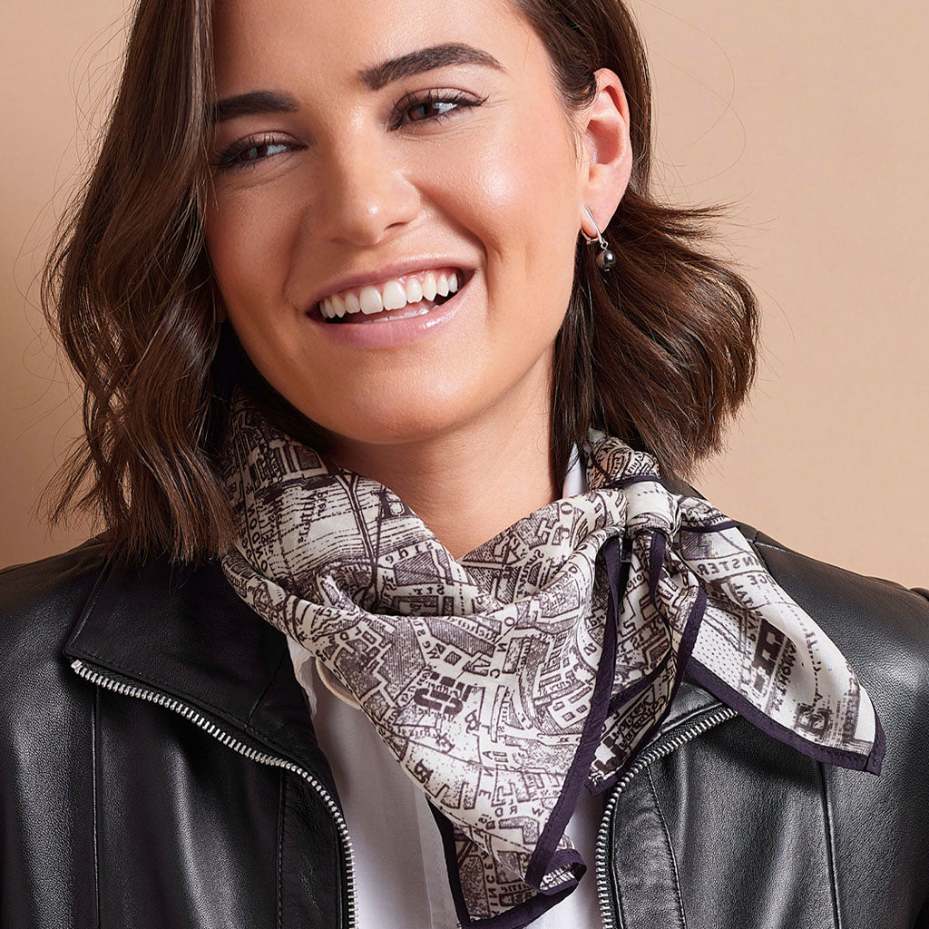 A woman with brown hair smiling while wearing a black leather jacket and a patterned scarf with a black and white city map design, set against a solid beige background.