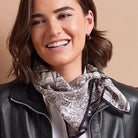 A woman with brown hair smiling while wearing a black leather jacket and a patterned scarf with a black and white city map design, set against a solid beige background.