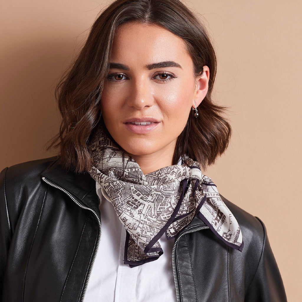 A woman with shoulder-length brown hair models a square scarf with a black and white architectural map pattern, styled around her neck over a white shirt and black leather jacket against a solid beige background.