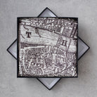 A square scarf with a black and white vintage map print, presented in a black box with a geometric star-shaped base on a gray textured background.