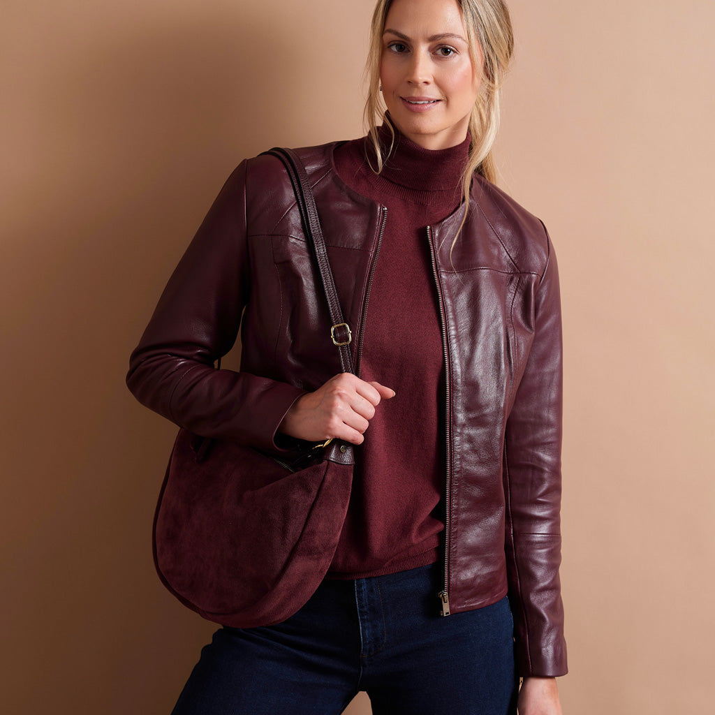 A woman wearing a dark burgundy leather moto jacket over a matching burgundy mock neck sweater and dark blue jeans, carrying a matching burgundy suede shoulder bag against a solid tan studio background.