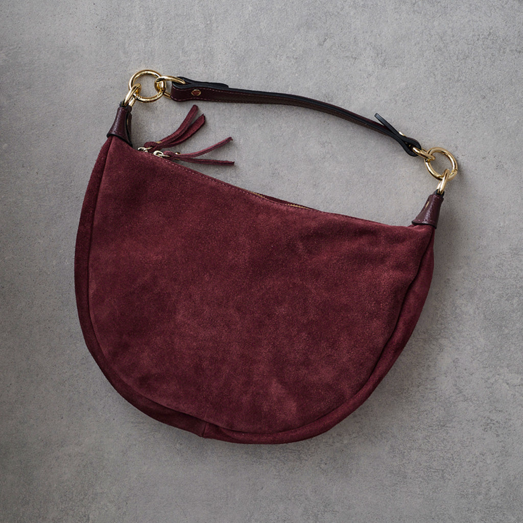 A burgundy suede crescent-shaped shoulder bag with a dark brown leather strap and gold-tone hardware, laid flat on a textured gray background.