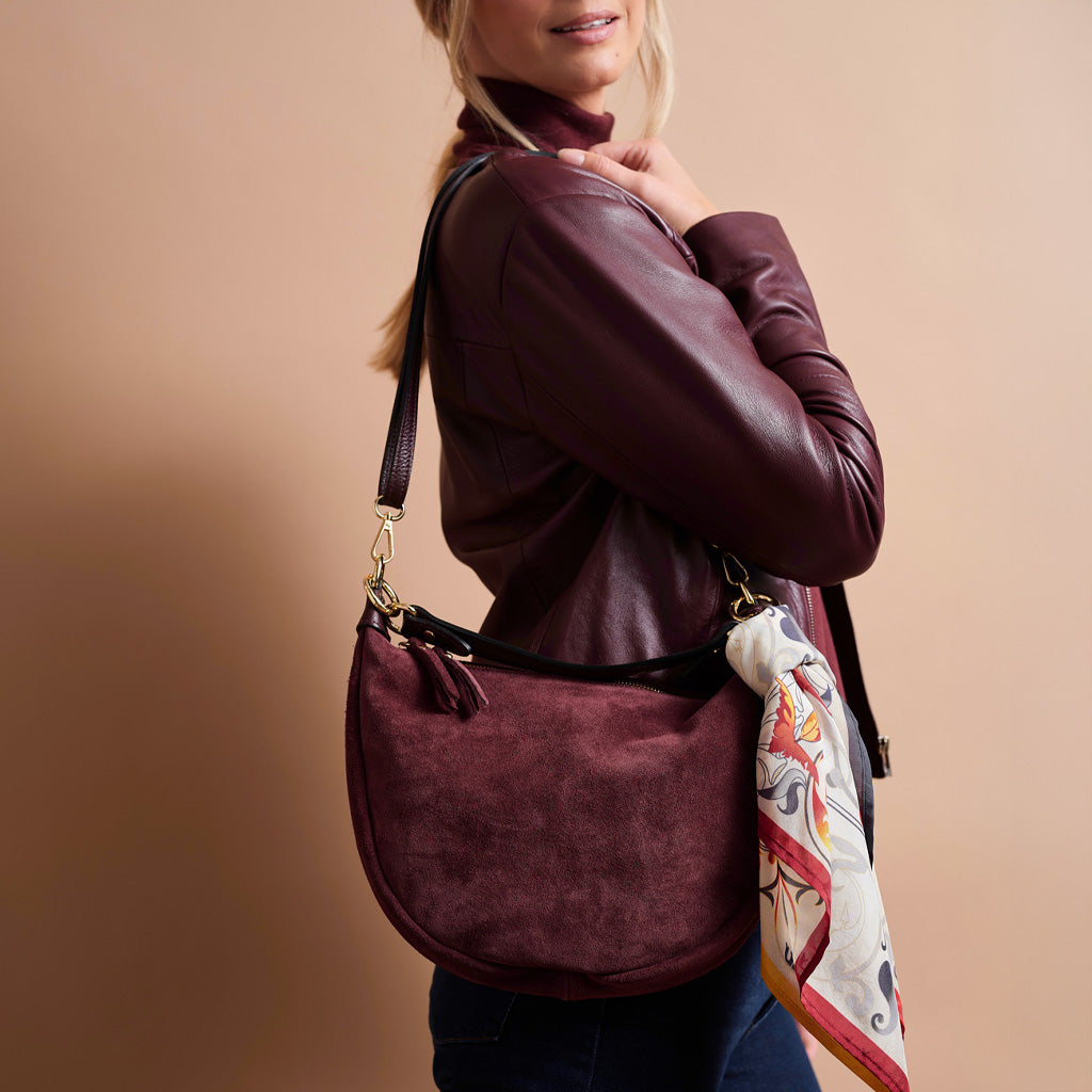 A woman wearing a burgundy leather jacket and dark blue jeans carries a matching burgundy suede shoulder bag with a patterned scarf draped over it, set against a solid beige background.