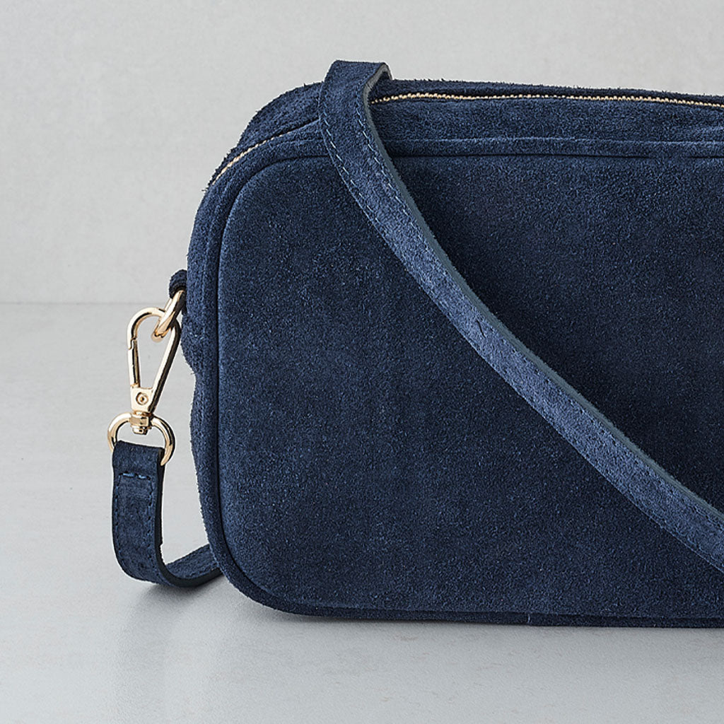 A navy blue suede crossbody bag with a matching fabric strap and gold-tone metal hardware, shown on a light gray background.