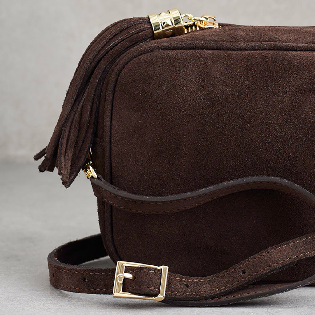 A close-up of a dark brown suede crossbody bag with a matching leather strap, gold-tone buckle, and a tassel zipper pull.