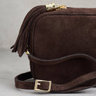 A close-up of a dark brown suede crossbody bag with a matching leather strap, gold-tone buckle, and a tassel zipper pull.