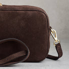 A close-up of a dark brown suede crossbody bag with a matching leather strap and gold-tone hardware, set against a neutral gray background.